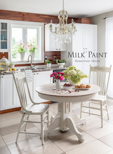 White kitchen featuring round table, pastries, flowers, chandelier; text reads: Limestone Milk Paint by Homestead House.