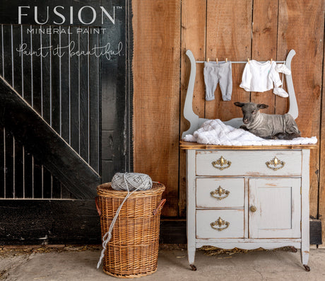Small dresser painted in Little Lamb by Fusion, with baby clothes inside and a wicker basket of yarn nearby.