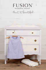 White vintage dresser with gold handles, painted in Fusion’s Little Stork, shown with lavender dress and blanket.