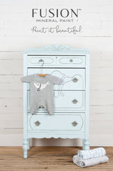 Dresser in Little Whale Fusion Mineral Paint with ornate handles, a onesie on top, and rolled blankets nearby.