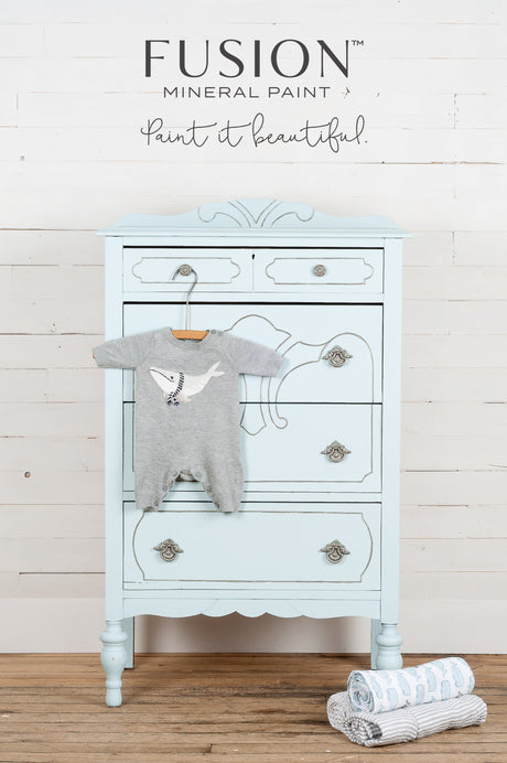 Dresser in Little Whale Fusion Mineral Paint with ornate handles, a onesie on top, and rolled blankets nearby.