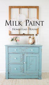 Blue cabinet with floral carvings, white knobs, and “Loyalist Milk Paint by Homestead House” above the clock.