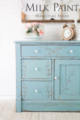 Distressed blue dresser with white knobs, floral carvings, and “Loyalist Milk Paint by Homestead House” text above.