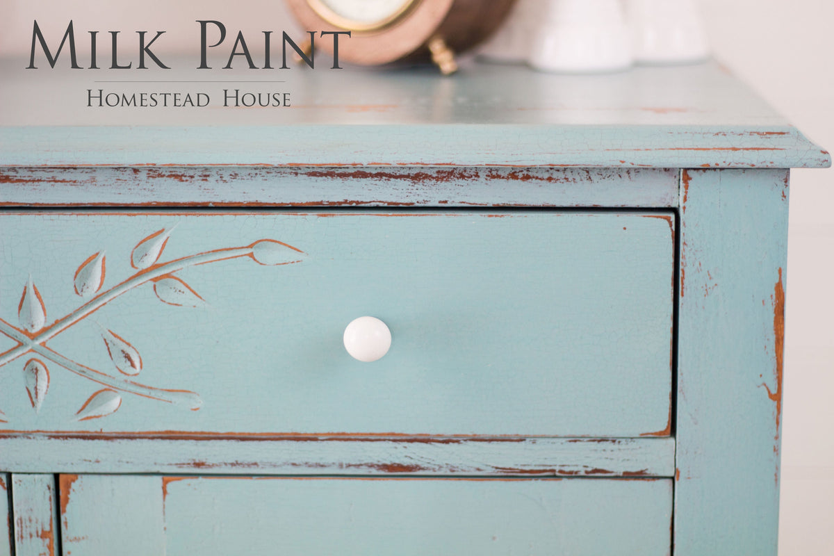 Distressed blue drawer with white knob, leaf design, and “Homestead House Loyalist Milk Paint” text in upper left.