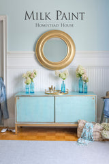 Light blue sideboard with vases, gold mirror, and Homestead House Loyalist Milk Paint text on the wall.