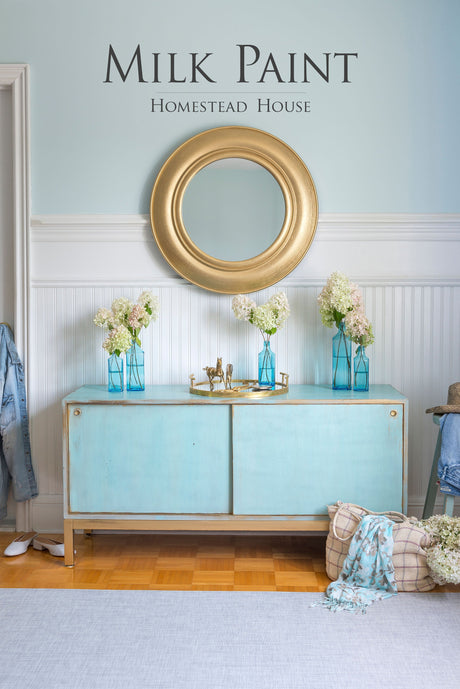Light blue sideboard with vases, gold mirror, and Homestead House Loyalist Milk Paint text on the wall.