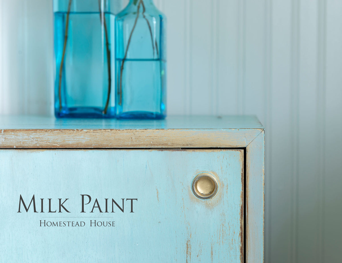 Light blue cabinet with brass knob, two blue bottles, and “Homestead House Loyalist Milk Paint” text on front.