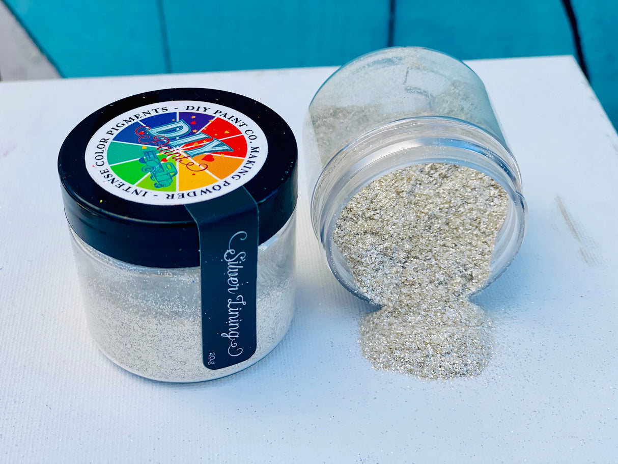 Two jars of Making Powder by DIY Paint Co., one tipped and spilling silver powder onto a white surface.