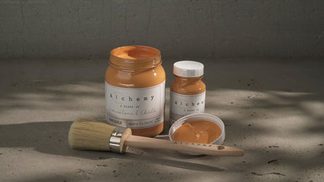 Two jars of Marigold Alchemy Paint by Fusion Mineral Paint with a paintbrush on a gray surface.