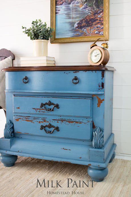 Distressed dresser in Homestead House Maritime Blue Milk Paint, styled with a plant, books, and a clock.