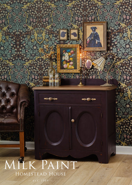 Dark wood cabinet in Homestead House Mennonite Red Milk Paint against floral wallpaper, near a leather chair.