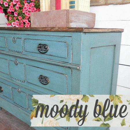Dresser painted in Old Fashioned Milk Paint Moody Blue with books, flowers, and ornate handles on top.
