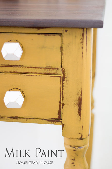 Distressed yellow table with two drawers, white knobs, and Mustard Milk Paint by Homestead House label.