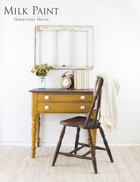 Yellow desk with white knobs painted in Mustard Milk Paint by Homestead House, shown with a chair, books, clock, and plant.