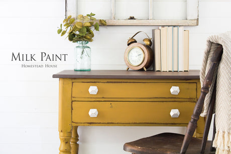 Yellow desk in Mustard Milk Paint by Homestead House with white knobs, books, a clock, and a leafy jar nearby.