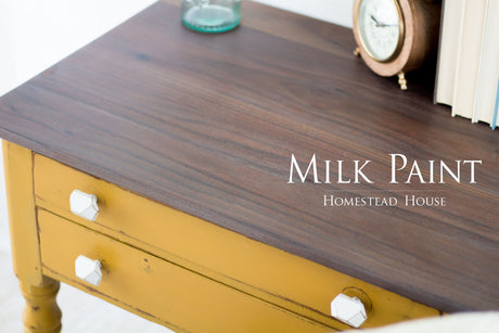Yellow wooden table painted Mustard Milk Paint by Homestead House, shown with clock, books, and glass bottle on top.