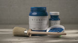 Paintbrush and two jars of Ocean Alchemy Paint by Fusion Mineral Paint on a textured surface in natural light.