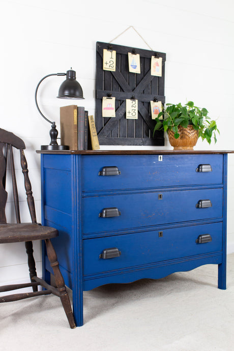 Dresser painted with Homestead House Ocean Milk Paint beside a chair, topped with lamp, plant, and artwork.
