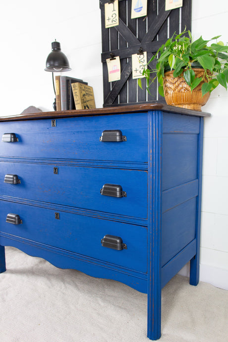 Dresser finished in Homestead House’s Ocean Milk Paint beside a black lamp, books, and basket holding a green plant.