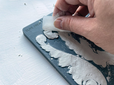 Hand lifting Olive Crest Mould by IOD from silicone mould, powder scattered on white surface.