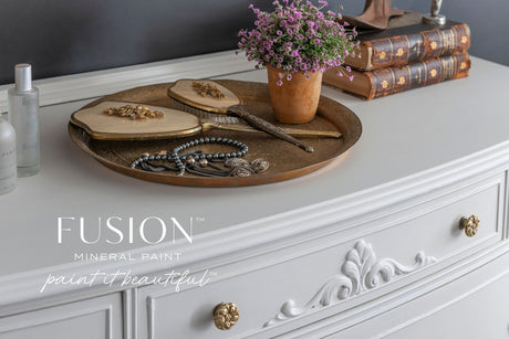 Dresser painted in Parchment Fusion Mineral Paint with gold knobs, topped with a brass tray, brushes, pearls, and books.
