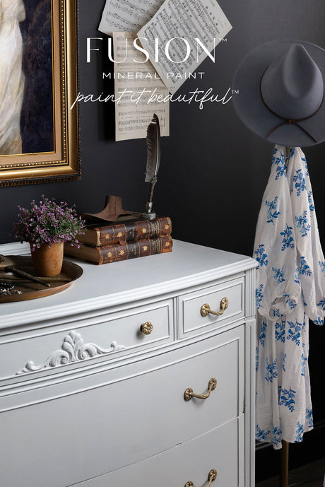 White dresser with brass handles, painted in Parchment by Fusion Mineral Paint, shown with vintage books and flowers.