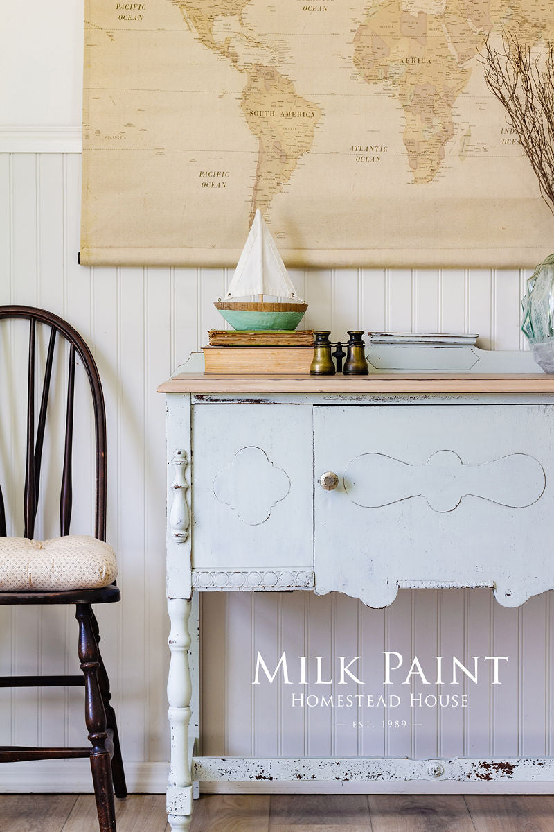 Side table finished in Homestead House Parlour Milk Paint, with sailboat model, books, and wall map; chair beside.