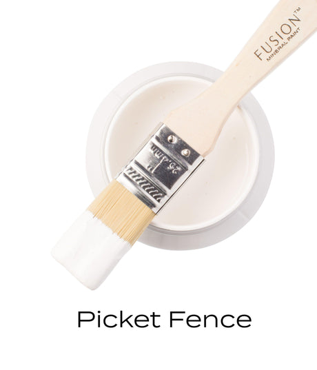 Brush with white paint rests on open Fusion Mineral Paint jar labeled “Picket Fence.”.
