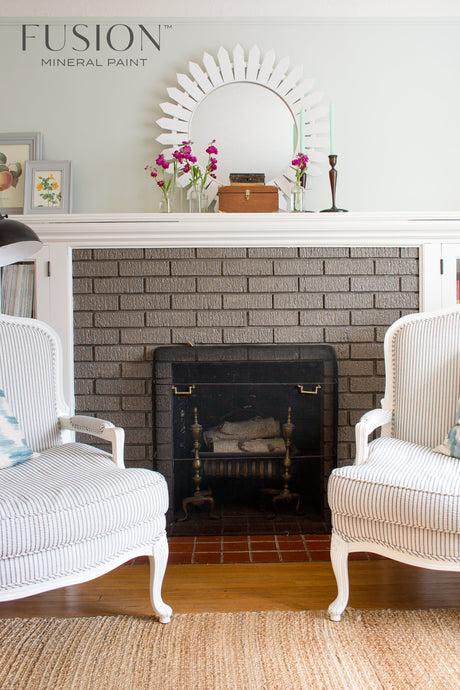 Brick fireplace with Picket Fence Fusion Mineral Paint mantel, two armchairs, flowers, and a round mirror above.