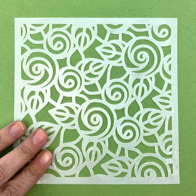 Hand holding floral square stencil for Notions Pixie Spray adhesive on green surface.