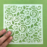 Hand holding floral square stencil for Notions Pixie Spray adhesive on green surface.