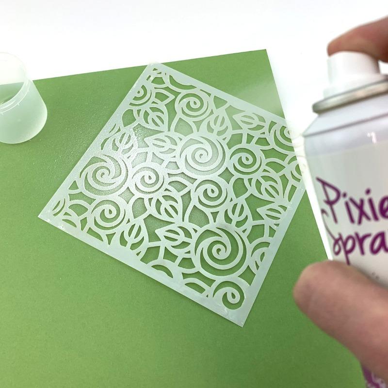 Hand spraying Notions Pixie Spray on a swirly leaf stencil placed on bright green paper.