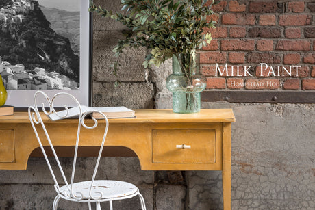 Yellow wooden desk with vase and book, white chair, and Prairie Sunset Milk Paint on brick wall.