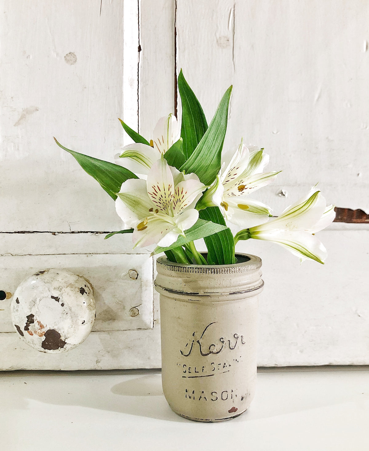 White lilies with green leaves arranged in a mason jar painted Proper by Old Fashioned Milk Paint, on a white cabinet.