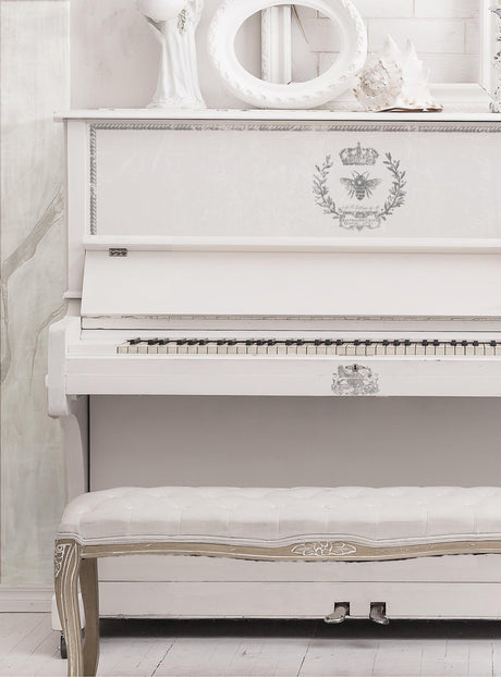 White upright piano with I.O.D. Queen Bee Stamp motifs, matching bench, and white decor on top.