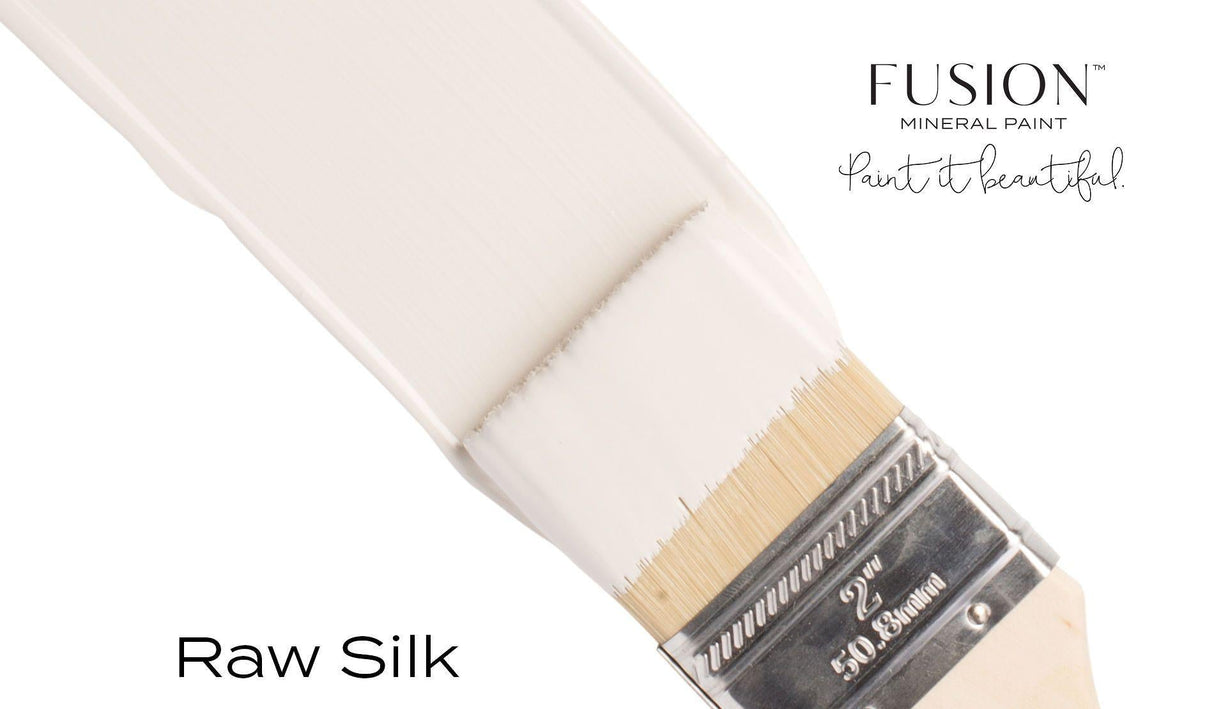 Paintbrush applying Fusion Mineral Paint Raw Silk acrylic to a white surface.