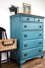 Teal dresser painted with Homestead House Renfrew Blue Milk Paint next to a dark chair in a bright room.