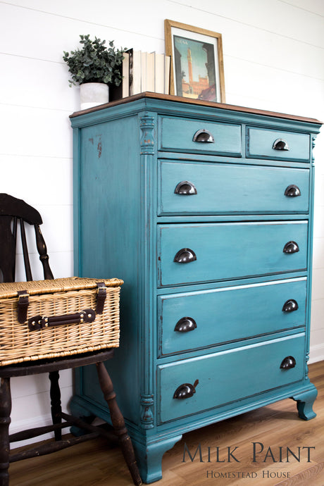 Teal dresser painted with Homestead House Renfrew Blue Milk Paint next to a dark chair in a bright room.