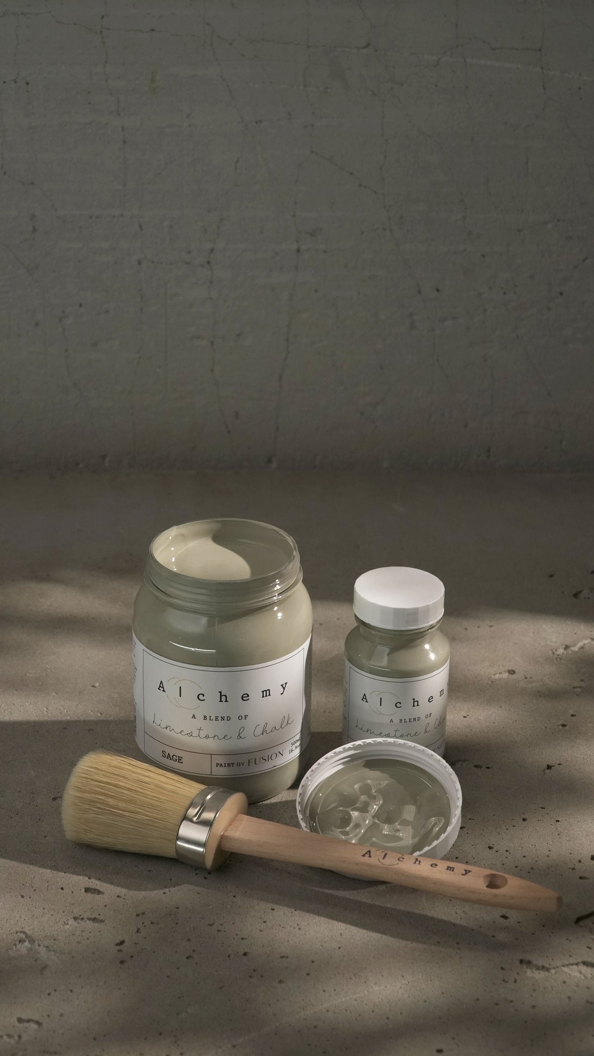 Three jars of Sage Alchemy Paint by Fusion Mineral Paint with a brush on a gray cracked surface.