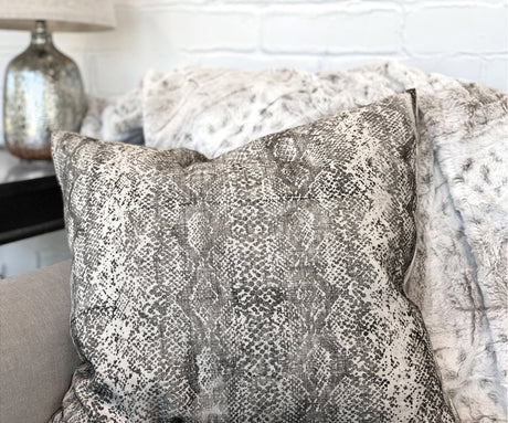 Pillow with retired Serpentine Stamp by I.O.D. displayed on a light sofa alongside a furry throw and metallic lamp.