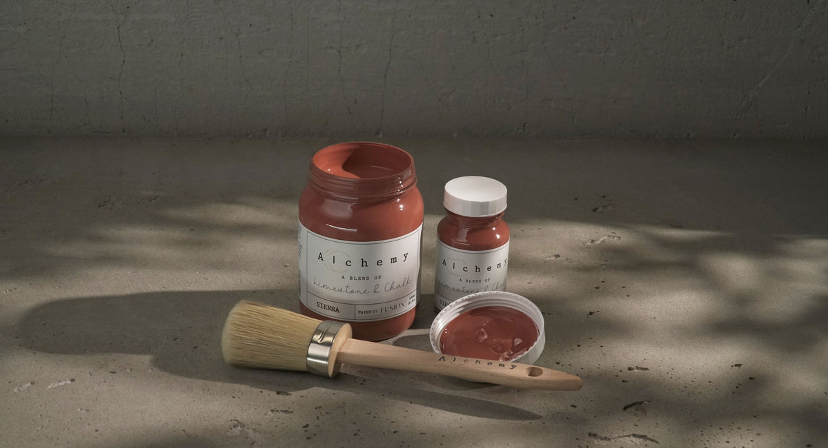 Three jars of Sienna Alchemy Fusion Mineral Paint and a brush on a textured surface in natural light.