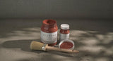 Three jars of Sienna Alchemy Fusion Mineral Paint and a brush on a textured surface in natural light.