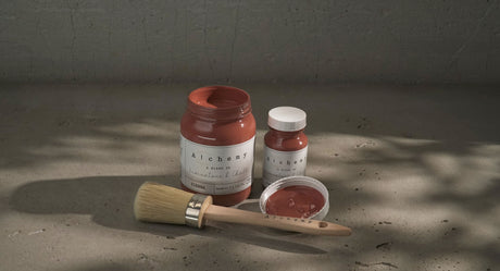Three jars of Sienna Alchemy Fusion Mineral Paint and a brush on a textured surface in natural light.