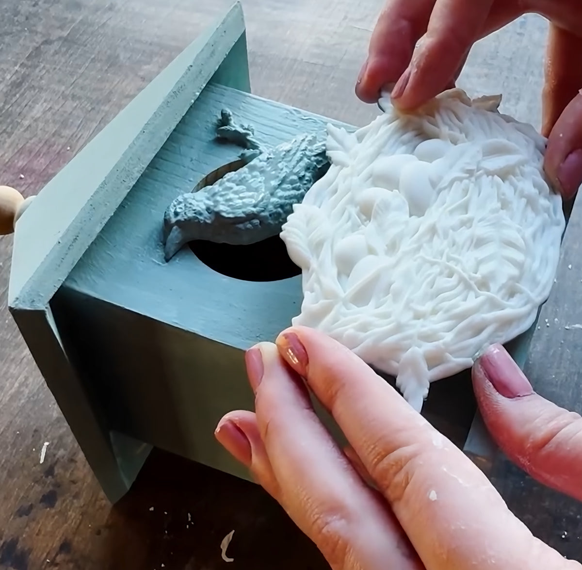 Hands holding a white Sparrow’s Nest Mould by I.O.D. beside a blue one, both on a wooden box.