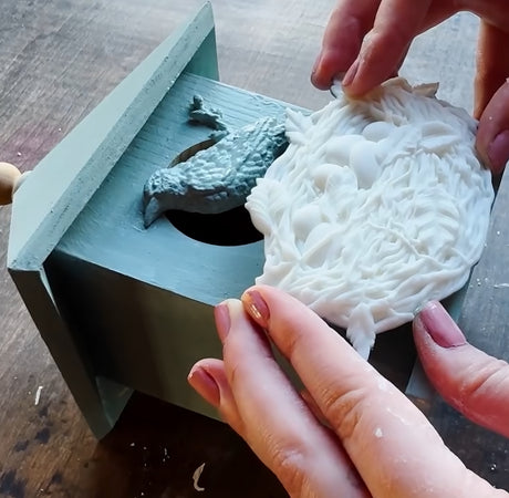 Hands holding a white Sparrow’s Nest Mould by I.O.D. beside a blue one, both on a wooden box.