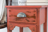 Red wood side table painted in Homestead House Spice Milk Paint with metal handle, books stacked on top, text above.