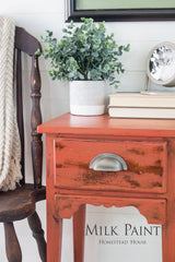 Orange side table painted with Homestead House Spice Milk Paint, shown with books, plant, clock, and chair.
