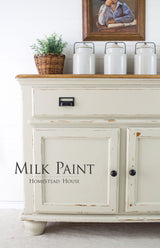 Cream distressed cabinet with jars, plant basket, and art. Text: Stone Fence Milk Paint by Homestead House.