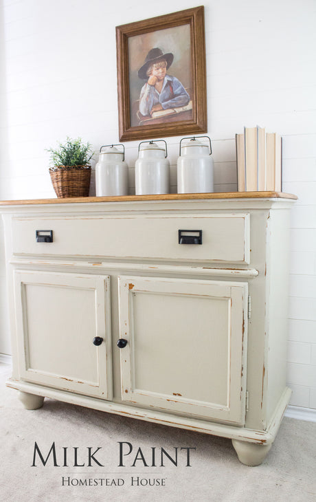 Distressed cream cabinet with jars, books, and “Stone Fence Milk Paint by Homestead House” label above a portrait.