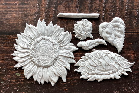 Six white clay I.O.D. Sunflowers Mould pieces and a leaf on a dark wood surface.
